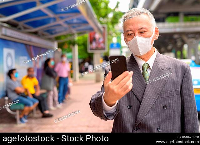 Portrait of mature Japanese businessman with mask for protection from corona virus outbreak at the bus stop in the city