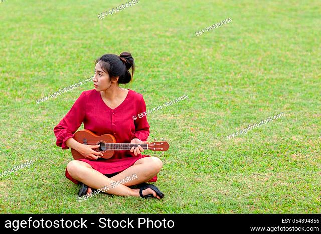 Portrait of beautiful young asian woman with red dress is playing ukulee with feeling happy and smile on green field at the public park