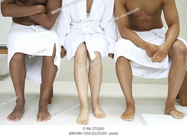 Two men in towels and woman in bathrobe sitting on bench in bathroom