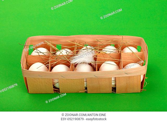 Ten fresh white farm chicken eggs in wooden container box with hay and feather over green background, high angle view