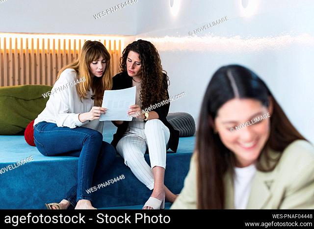 Two businesswomen reading a document in office lounge with colleague in foreground