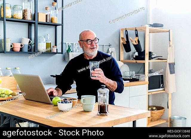 Senior man with laptop holding drinking glass at home
