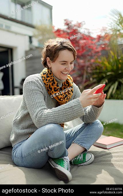 Happy young woman using smart phone on sunny patio lounge chair