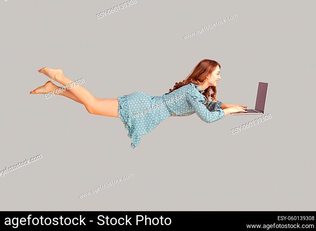 Hovering in air. Happy cheerful girl ruffle dress levitating with laptop, typing keyboard, reading good news message on computer while flying in mid-air