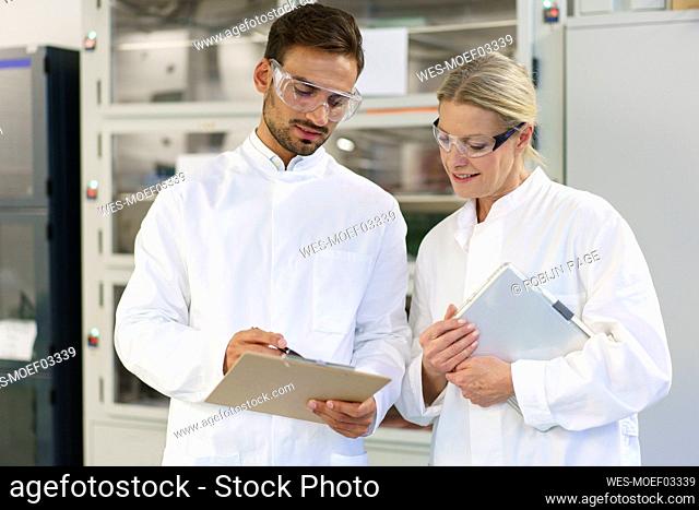 Young male technician discussing over clipboard with female blond colleague holding digital tablet at laboratory