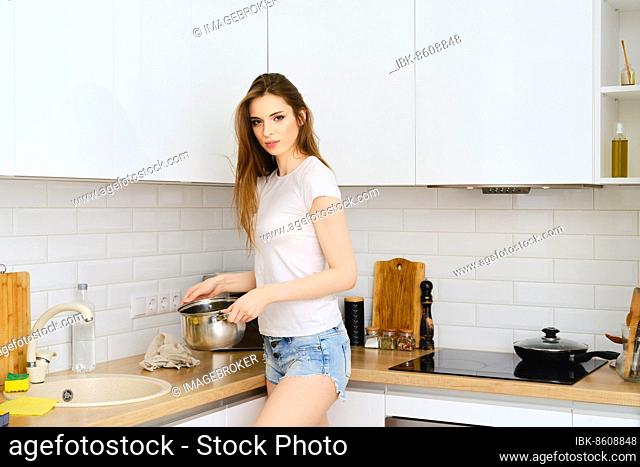 Young attractive woman pouring pure water to stainless steel stockpot. She is going to put it on electric stove. Beautiful female preparing oatmeal porridge for...