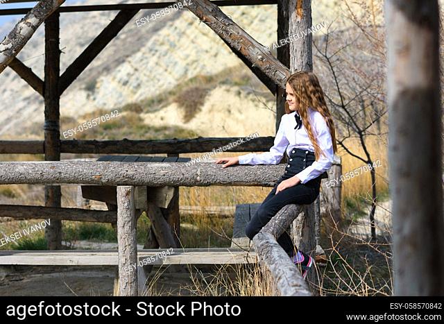 A girl sits on a wooden fence near an old gazebo made of wooden beams on a sunny warm day
