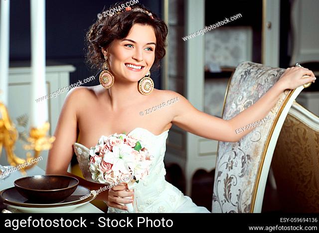Beautiful young bride in fashionable gown with floral bouquet sitting near dinner table. Interior photo