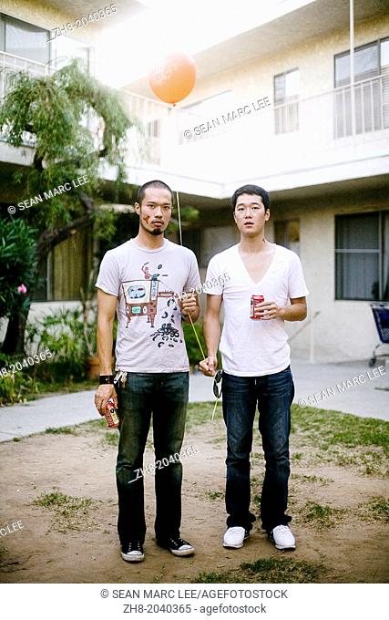 Two Asian men holding beers and a balloon in a courtyard in an apartment complex in Los Angeles