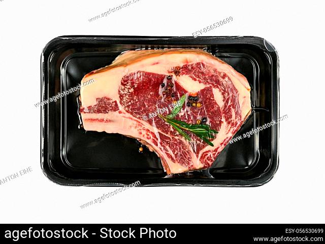 Close up one marbled raw ribeye beef steak with rib bone vacuum sealed with black plastic film, isolated on white background, elevated top view, directly above