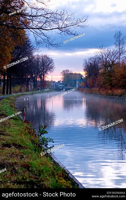 River stream canal bending with grass banks and wild flowers and trees in a scenic landscape on a misty autumns morning sunrise day
