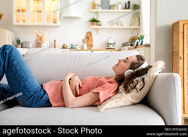Happy woman lying on sofa listening music through wireless headphones