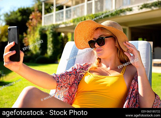 Caucasian woman sitting in sunny garden wearing sunhat and sunglasses taking selfie with smartphone