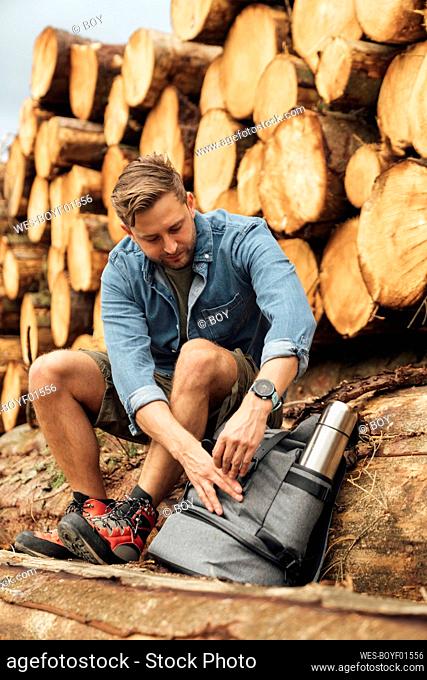 Male hiker with backpack sitting on log against woodpile in forest