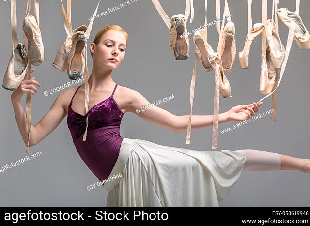 Pretty ballerina in a violet top and a cream skirt posing on the gray background in the studio. Around her there are many hanging beige pointe shoes