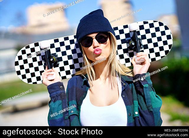 Young woman in knit hat puckering while carrying skateboard on shoulder during sunny day