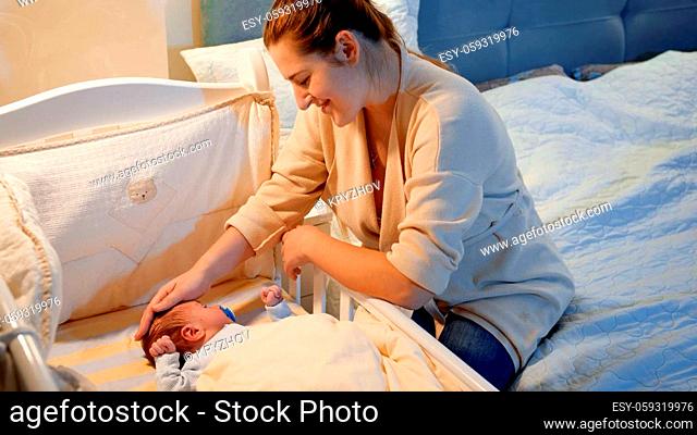 Happy smiling mother lookig on and stroking her little baby son lying in crib at during sleepless night. Concept of happy parenting and family happiness