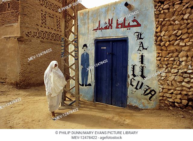A veiled muslim woman walks past a door with Charlie Chaplin sign on the wall, Nefta, Tunisia