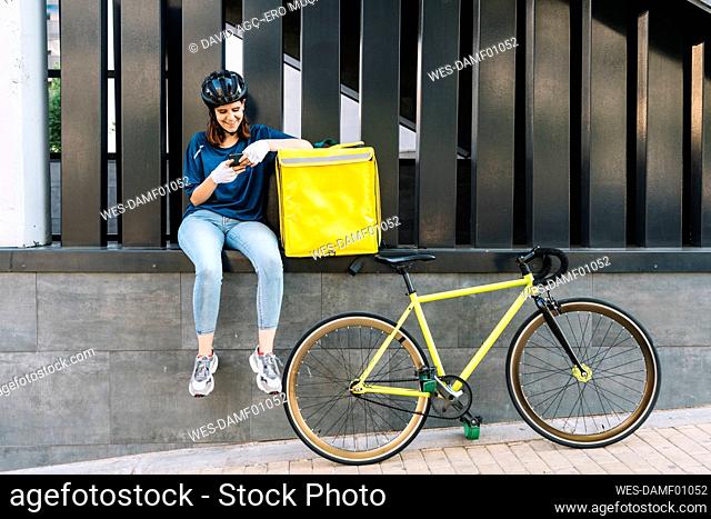 Smiling delivery woman using smart phone sitting on wall