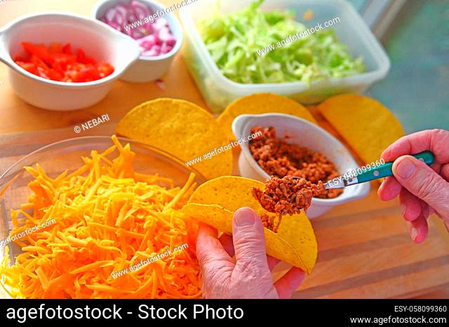 A man makes tacos at home with taco shells, ground beef, lettuce, onions, tomatoes and grated cheese