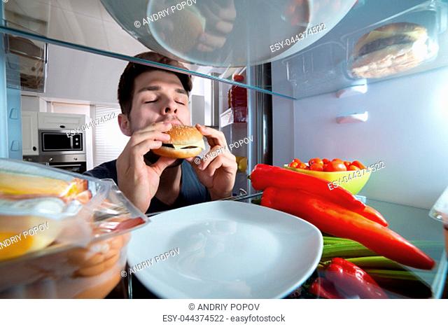 Close-up Of A Hungry Young Man Eating Burger Near Refrigerator