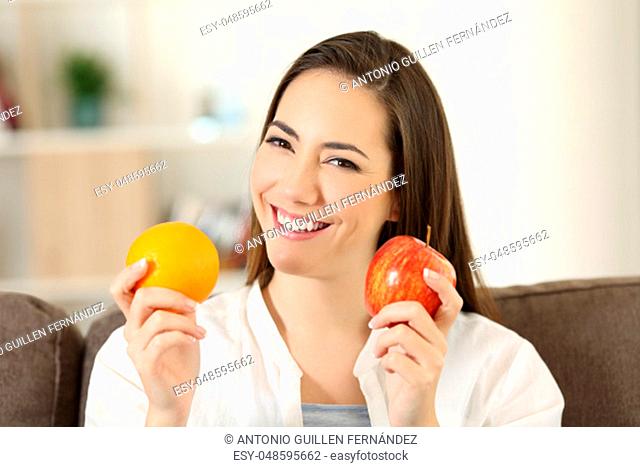 Front view portrait of a happy woman holding an apple and orange looking at camera sitting on a couch in the living room at home