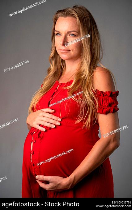 Pretty pregnant woman in red dress holding her belly while standing over gray wall background. Maternity concept