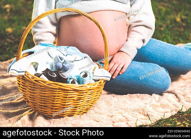 Pregnant woman expecting a baby, posing outdoors with a wicker basket with clothes for her newborn