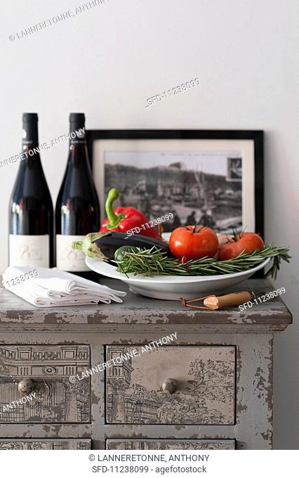 A plate of vegetables with rosemary, bottles of red wine and napkins on a chest of drawers