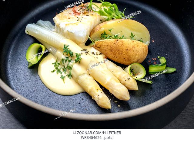 Modern German fried cod fish filet with white asparagus in hollandaise sauce und roast potatoes as close up on a plate