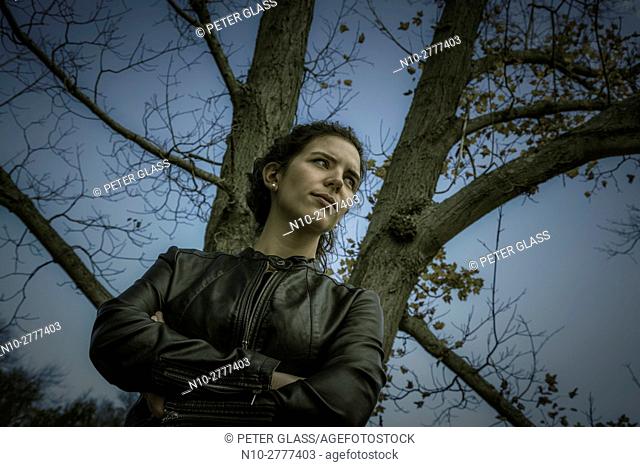 Young woman in a leather jacket standing next to a tree with no leaves