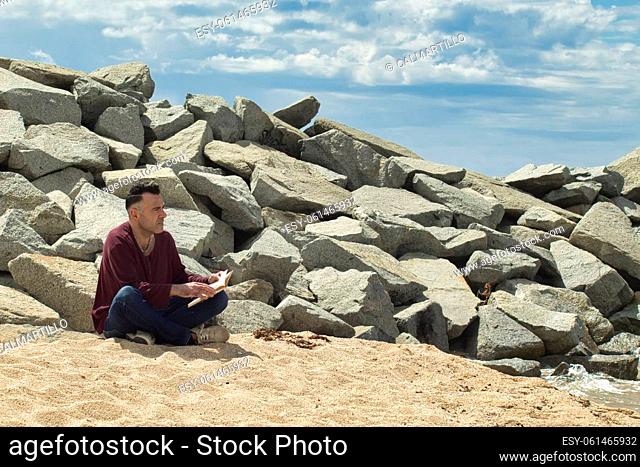 man sitting on the sand reading a book on the seashore near a breakwater