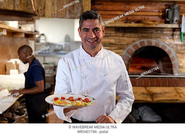 Portrait close up of a smiling middle aged Caucasian male chef holding a dish of prepared food in a restaurant kitchen, while a male member of kitchen staff...