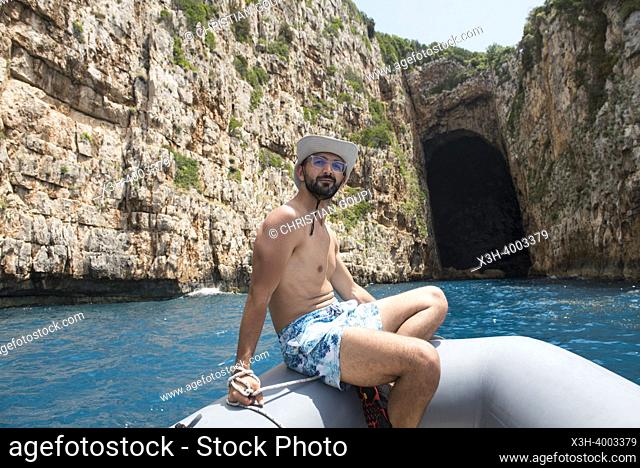 Shirtless man sitting at the bow of a zodiac in front of the Haxhi Ali cave, Peninsula of Karaburun, within the Karaburun-Sazan Marine Parc, Vlore bay, Albania