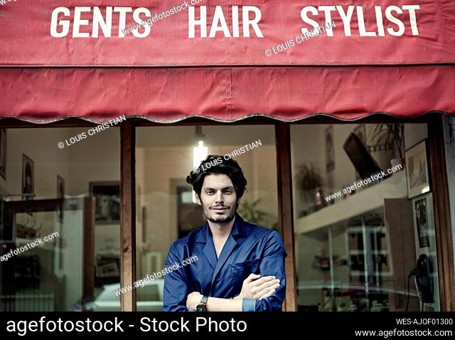 Portrait of handsome barber standing in front of barber shop with crossed arms