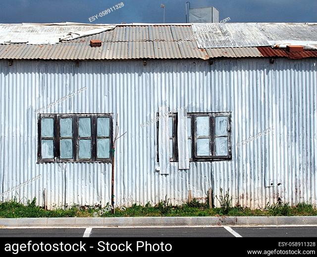 full frame close up of a shabby old dilapidated corrugated iron building with with closed painted over patched windows repaired rusting roof and white faded...