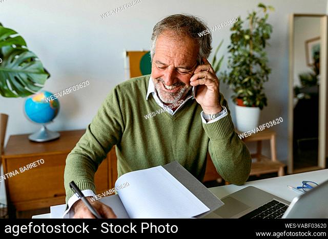 Smiling man writing in book while talking on mobile phone at home