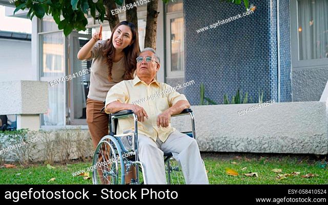 Disabled senior man on wheelchair with daughter, Happy Asian generation family having fun together outdoors backyard, Care helper young woman walking an elderly...