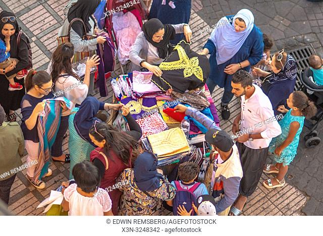 High angle view of shoppers browsing colorful djellaba and other textile products at outdoor marketplace in Jemaa el-Fnaa square, Marrekech, Morocco
