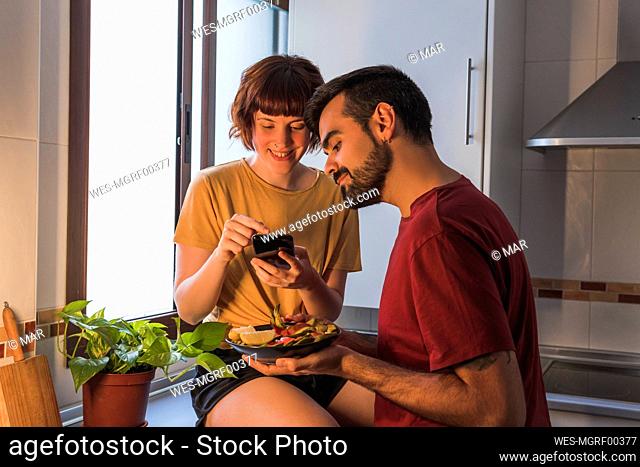 Smiling woman photographing food held by boyfriend in kitchen at home