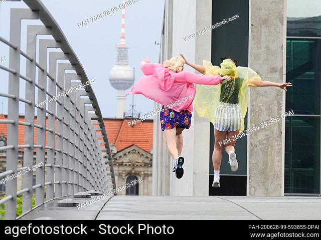 28 June 2020, Berlin: Two young women jump over the Marie-Elisabeth-Steg at Paul-Lübbe Haus after a downpour of rain with colourful rain capes