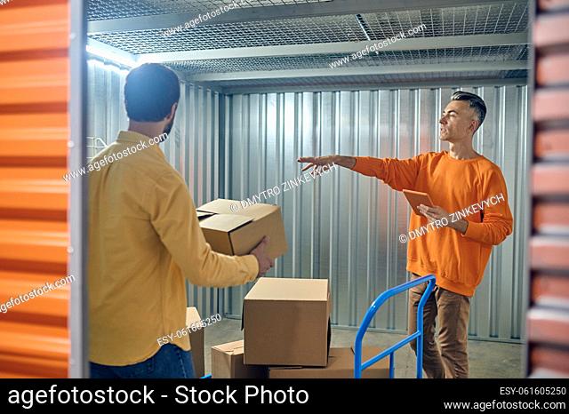 Warehouse manager with the tablet computer pointing at the rack to the worker with the cardboard box
