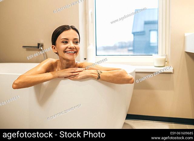Merry pretty woman leaning on the edge of the bath and looking into the distance