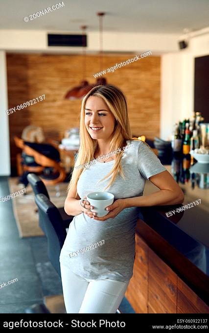 Smiling happy young pregnant woman relaxing leaning against a bar counter with a mug of hot tea cradling her baby bump in her hand as she bonds with her unborn...
