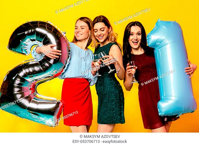 Birthday party. Three amazing attractive women in trendy dressed celebrating an anniversary with champagne. Isolated over yellow background