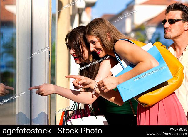 Two women being friends shopping downtown with colorful shopping bags, they are lolling into a glass store door and are amazed; in the background a man is...