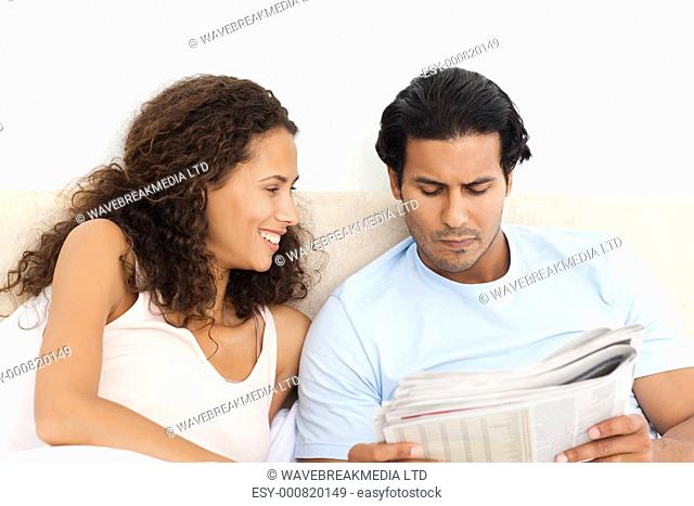 Happy woman looking at her serious boyfriend reading the newspaper on the bed