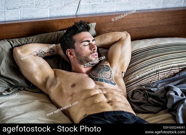 Shirtless sexy muscular male model lying alone on his bed in his bedroom, looking away with a seductive attitude