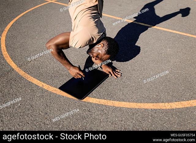 Man using graphics tablet while doing handstand on sports court during sunny day