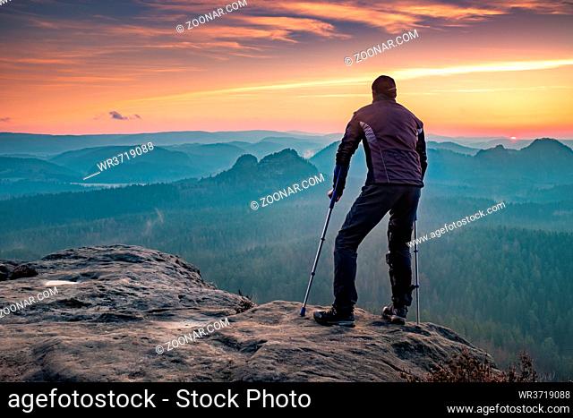 Disabled tourist with crutches. Rear view of Hurt hiker man with forearm poles walking against mountain sunset background